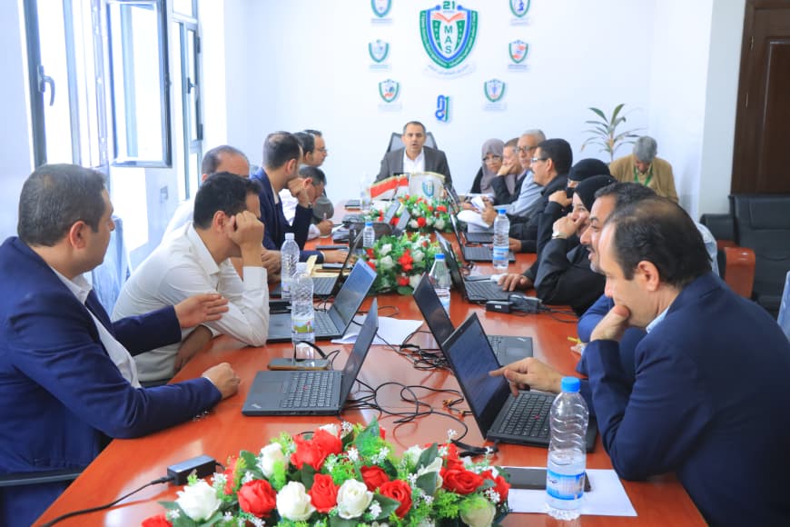 Group of professionals around a long conference table with laptops, discussing a meeting agenda in a bright office.