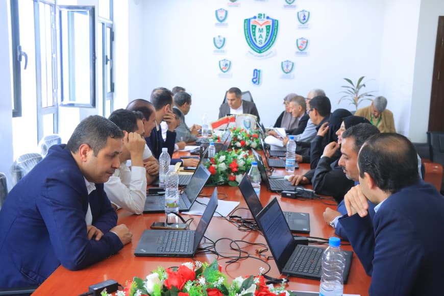 Group of professionals seated around a long conference table with laptops and water bottles in a bright meeting room with a crest on the wall behind them.