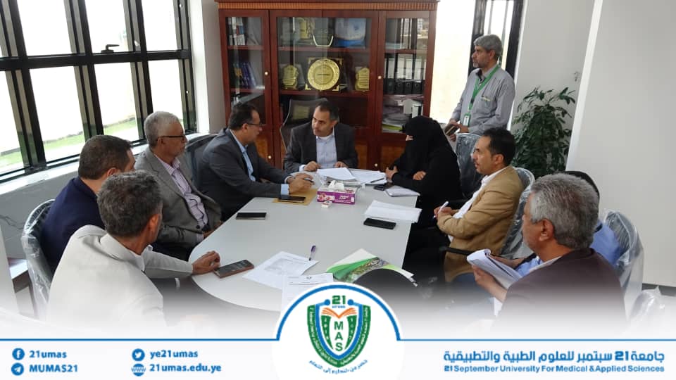 Group of professionals seated around a conference table in discussion, with documents and laptops visible, in an office with a trophy cabinet in the background, banner at bottom says university logo and staff.
