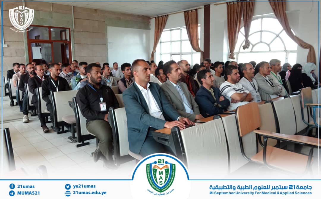 Audience of adults seated in a university lecture hall attending a seminar, with attendees wearing ID badges and a university banner visible in the foreground.