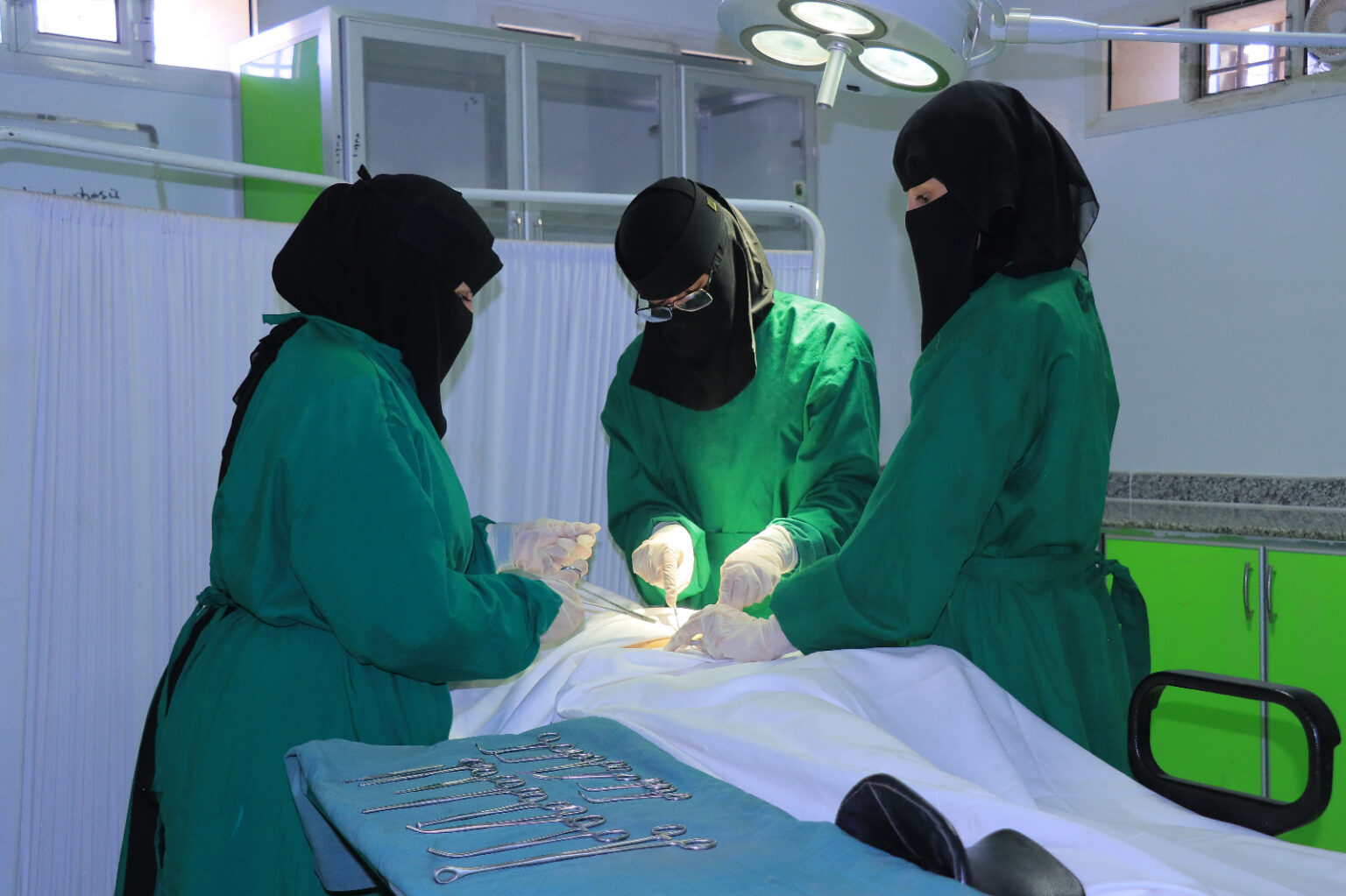 Three surgeons in green gowns and black head coverings operate on a patient in an operating room, with surgical tools laid out on a table nearby.