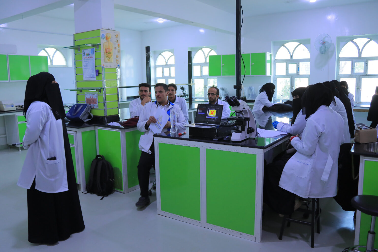 Group of students in white lab coats in a bright science lab with green cabinets and a microscope on the bench.