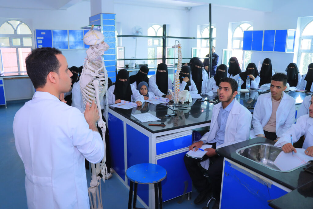 Instructor in a white lab coat explains a human skeleton to a classroom of students in a blue-tiled science lab with skeleton models on stands.