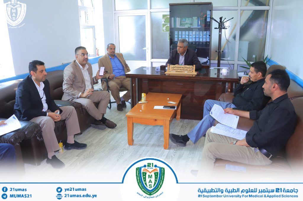 IMG-20260831-WA0040 - جامعة 21 سبتمبر Six men sit in a semi-circle in a bright office, wearing business attire, talking around a wooden desk with a nameplate in Arabic. A university logo is visible at the bottom edge.