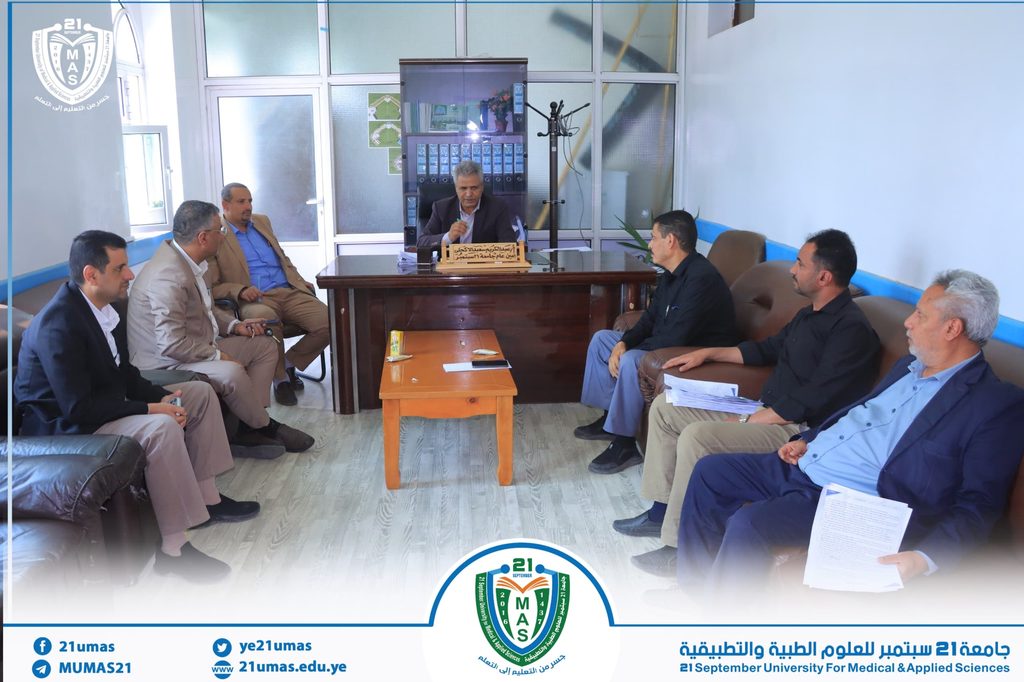 IMG-20260901-WA0005 - جامعة 21 سبتمبر Group of men seated around a dark wooden desk in a bright meeting room, central speaker addressing the others with a microphone present; MAS logo visible on the wall behind.