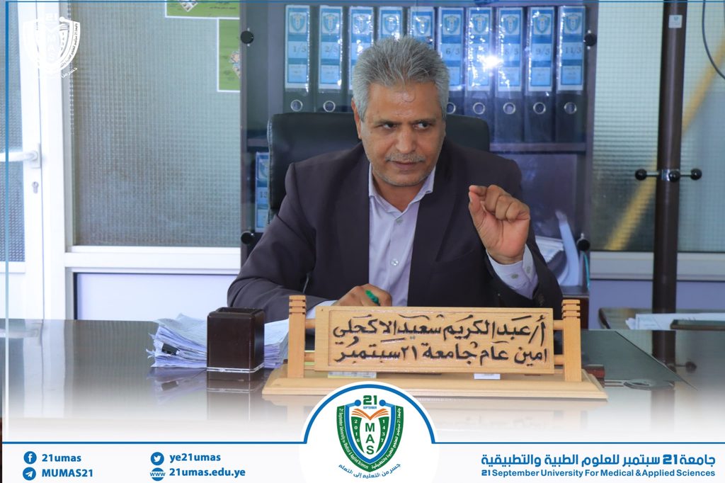 IMG-20260901-WA0007 - جامعة 21 سبتمبر Man in a suit sits at a desk in an office, writing with a green pen; a wooden Arabic nameplate is in front, with blue binders behind and a university banner below.
