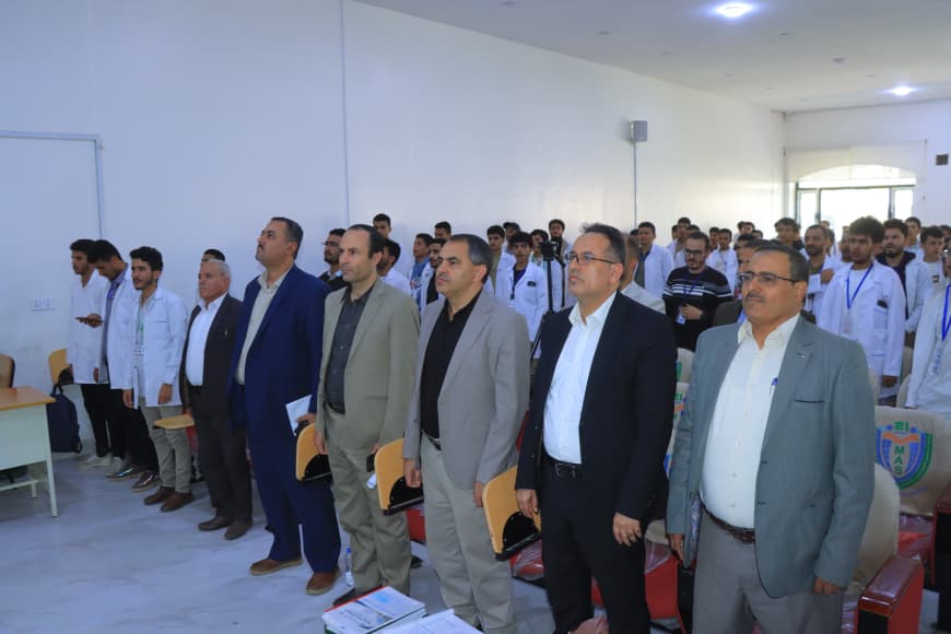 Group of formally dressed men standing at the front of a lecture hall while students in lab coats listen in the background.