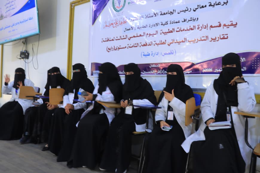 Group of women wearing white lab coats and black hijabs seated in a row in a conference room, holding notebooks and papers, with an Arabic banner on the wall behind them.