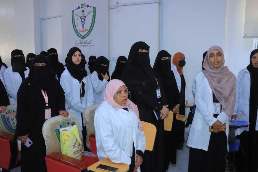 Group of female medical students in white coats standing in a classroom, many wearing hijabs or niqabs, with a medical emblem on the wall behind them.