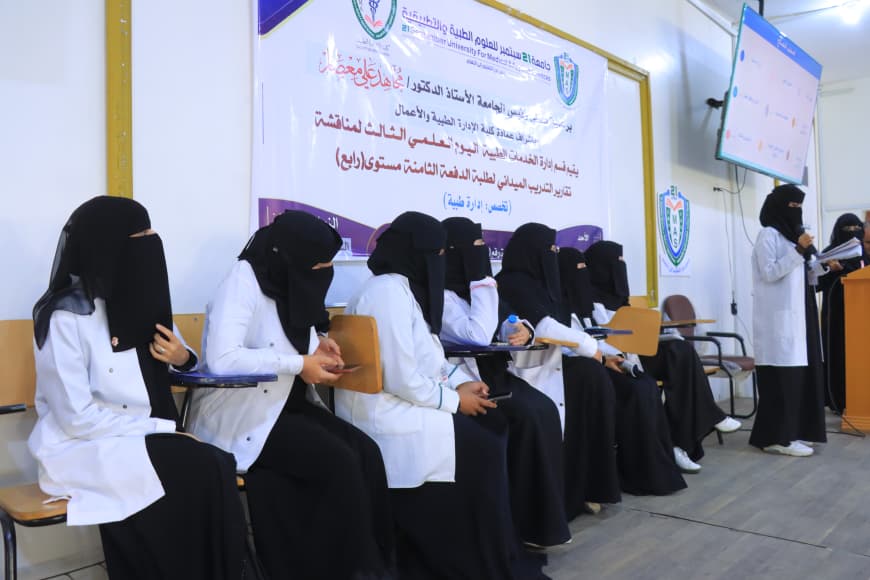Row of women in white coats and black hijabs seated in a conference room, with an Arabic banner behind them; a presenter stands at a podium on the right.