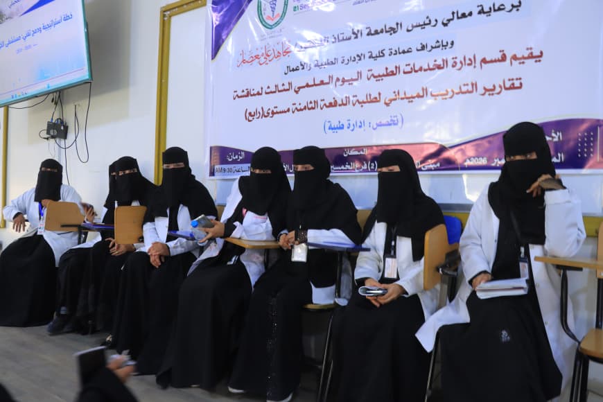 Group of women in black abayas and niqabs seated in a row, holding notebooks and pens during a workshop with an Arabic banner in the background.