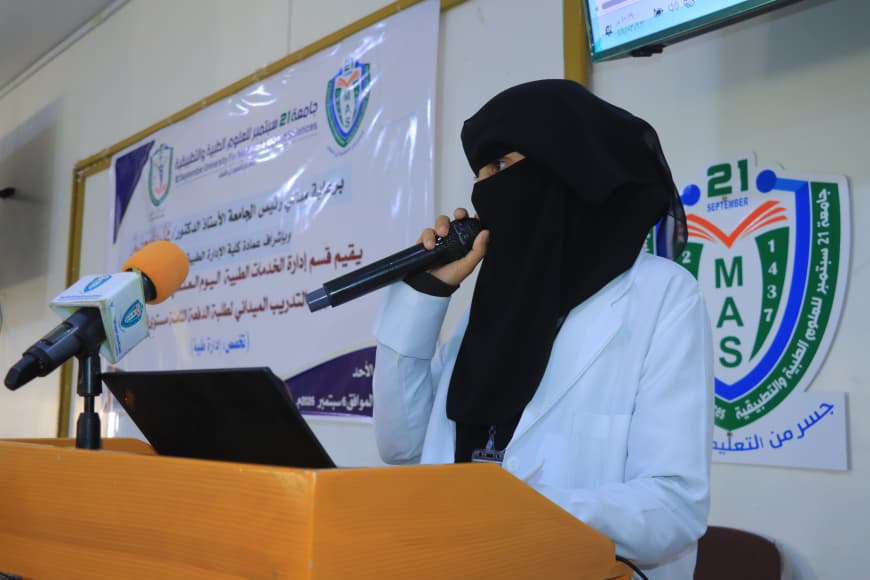 Female speaker wearing a black hijab and white coat presents at a wooden podium with a microphone and laptop, in front of a banner and MAS emblem.