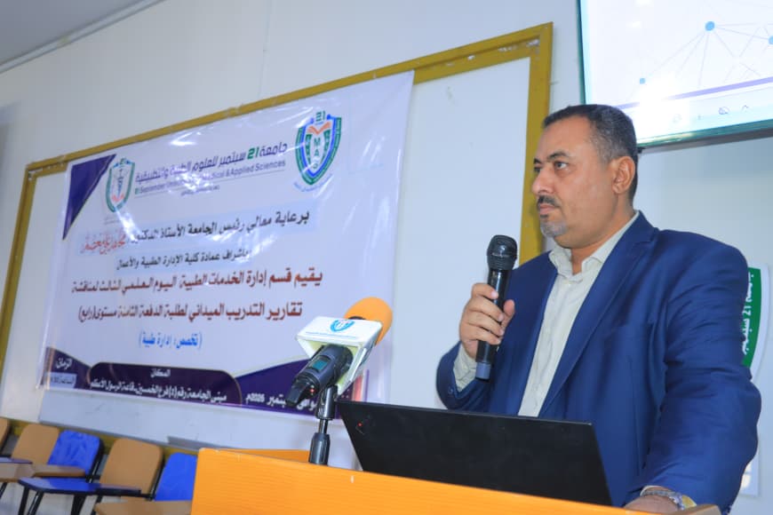 Man in a blue suit presenting at a podium with a microphone, banner with Arabic text in the background.
