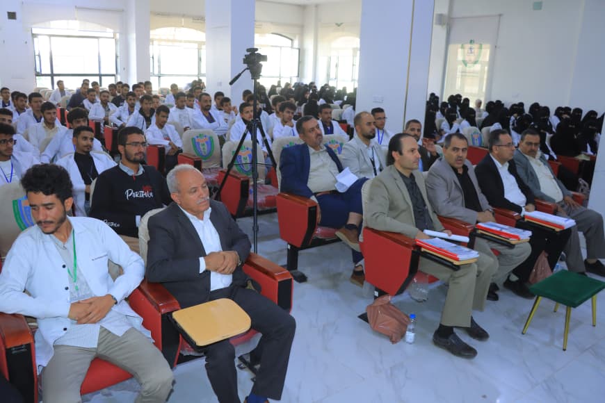 Audience in a bright conference hall; front row of men in suits with folders, behind them students in white shirts.