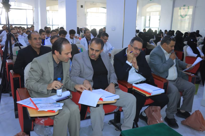 Four men in the front row of a conference reviewing papers and taking notes, with many attendees behind them.
