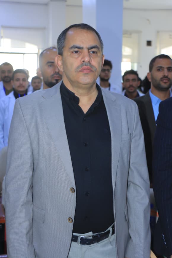 Man in a light gray blazer and black shirt stands at the front while a group watches in a hall in the background.