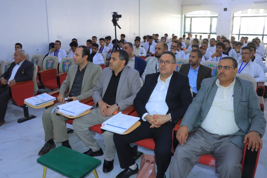 Front-row executives seated with folders and notebooks during a formal seminar; audience in matching shirts sits in rows behind them, a camera on a tripod records the event.