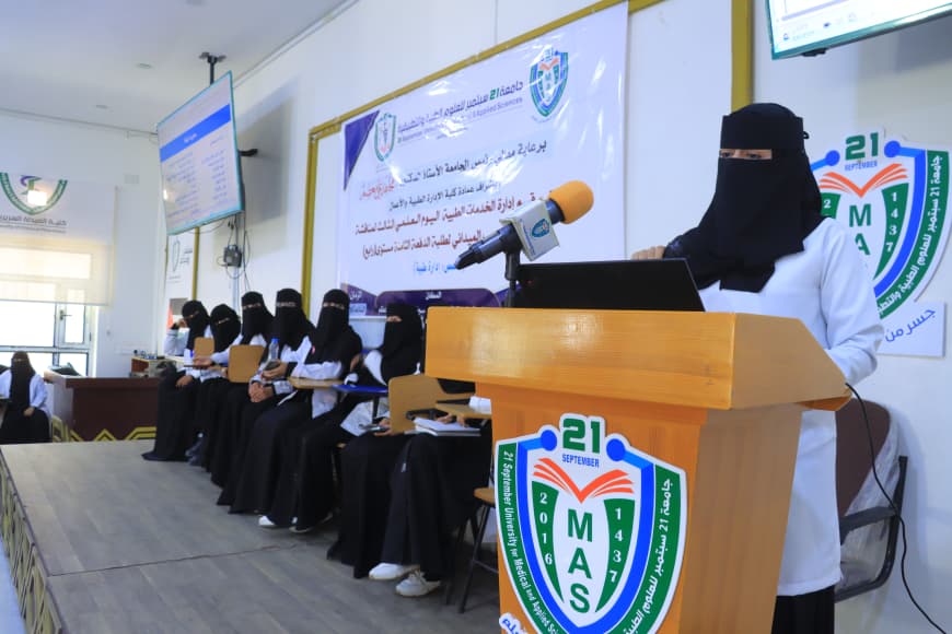 A woman wearing a black hijab and white coat speaks at a wooden podium with a laptop and microphone, while seated women in black abayas listen in a conference room with banners and a logo on display.