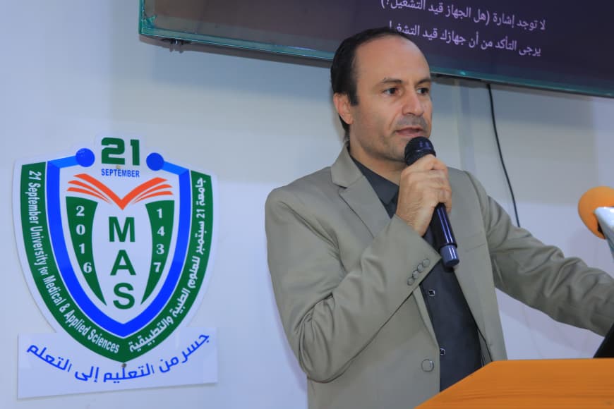 Man in a beige suit speaks at a podium with a microphone, standing beside a large MAS emblem and Arabic banner in a conference setting.