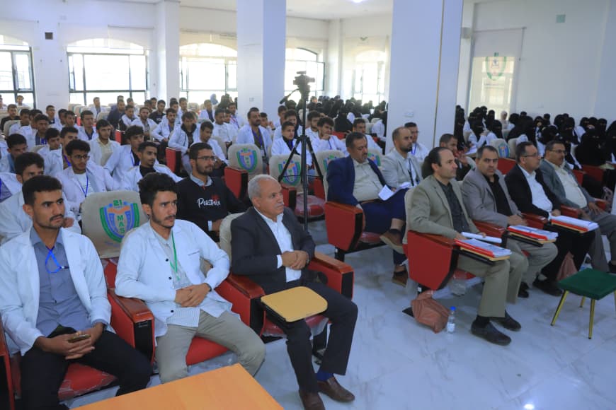 Lecture hall full of students in white coats listening to a presentation; front row hosts in suits with notebooks nearby on orange desks.