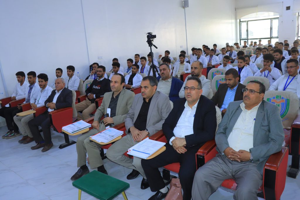 Large lecture hall filled with students, many in white shirts, seated in rows with notebooks; a video camera on a tripod stands center stage in the aisle.