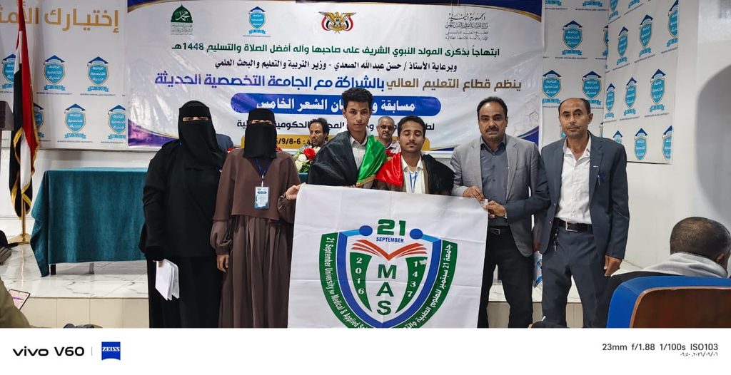Group of six people standing on a stage, including two women in full black abayas, posing with a white flag featuring a green crest and the letters 'MAS' and the number 21, in front of an Arabic banner and backdrop of event logos.