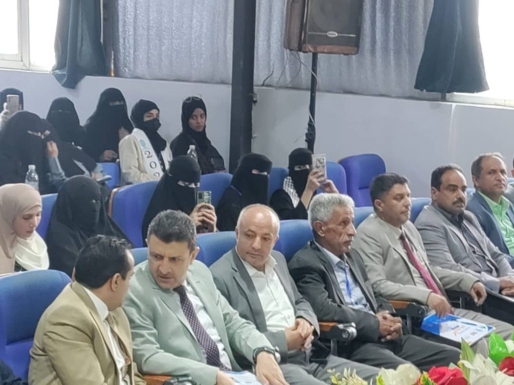 Row of men in suits seated in the front row of a conference hall, with audience members, including women in niqab, seated behind them and some taking photos with smartphones.
