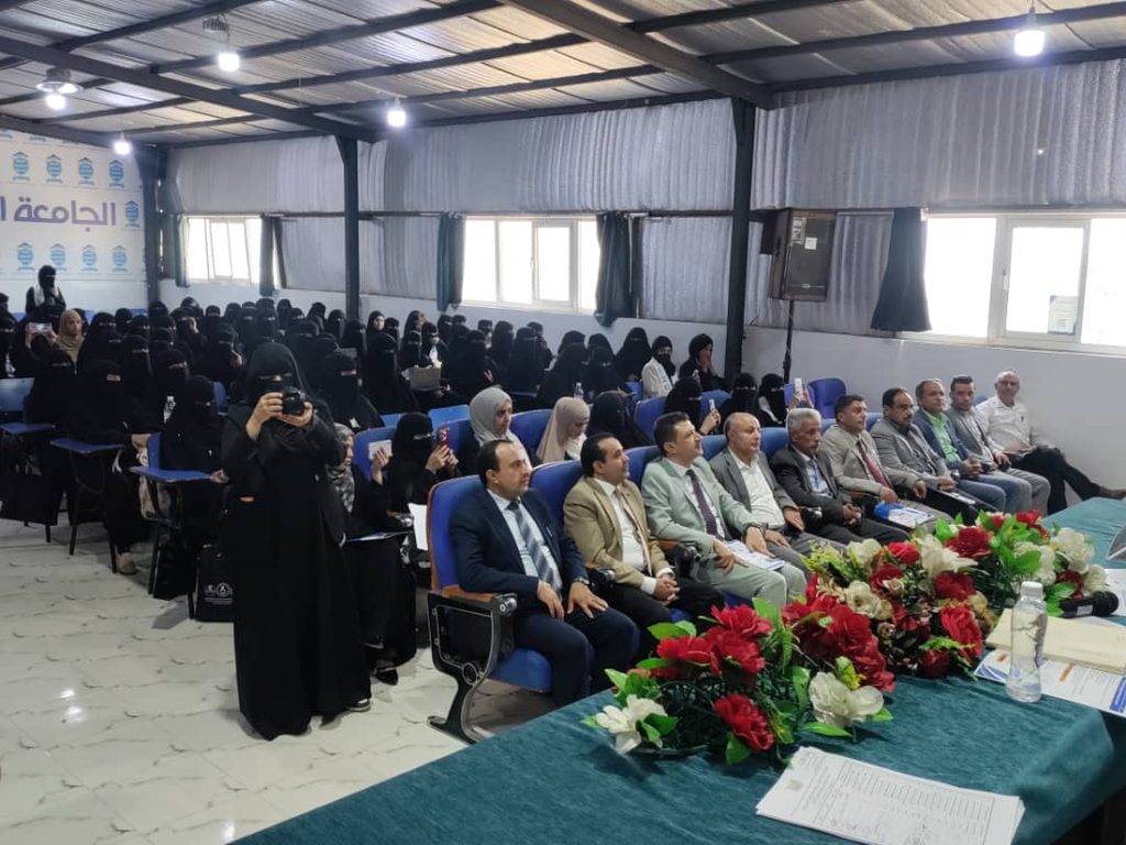 Front row of men in suits seated at a conference table with a large floral centerpiece, while many women wearing black abayas and niqabs sit in the audience; a photographer in black is taking a photo near the left side.