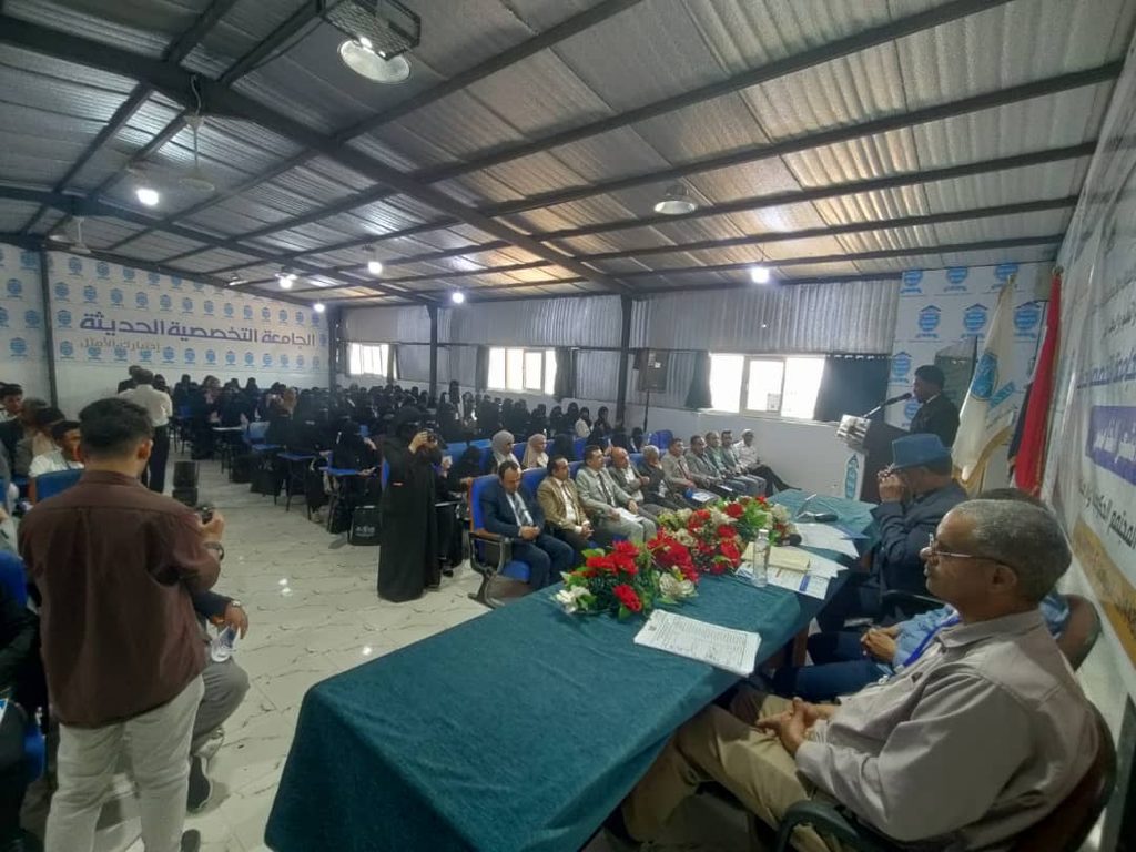 Panel of officials seated at a long table on a stage in a crowded conference hall, audience watching attentively.