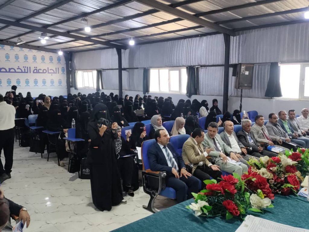 Large indoor conference with rows of attendees, many women in black abayas and men in suits, seated facing a panel at a decorated table with red and white flowers in the foreground.