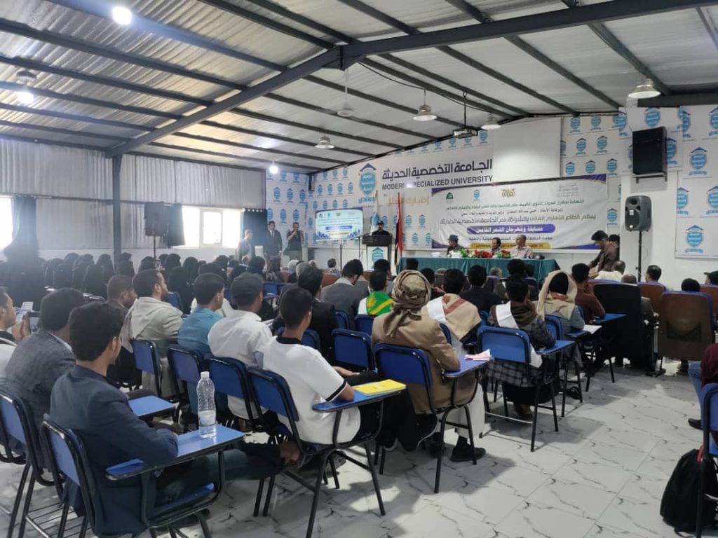 Audience seated in a conference hall listening to a panel on stage with a long banner in Arabic behind them at Modern Specialized University.