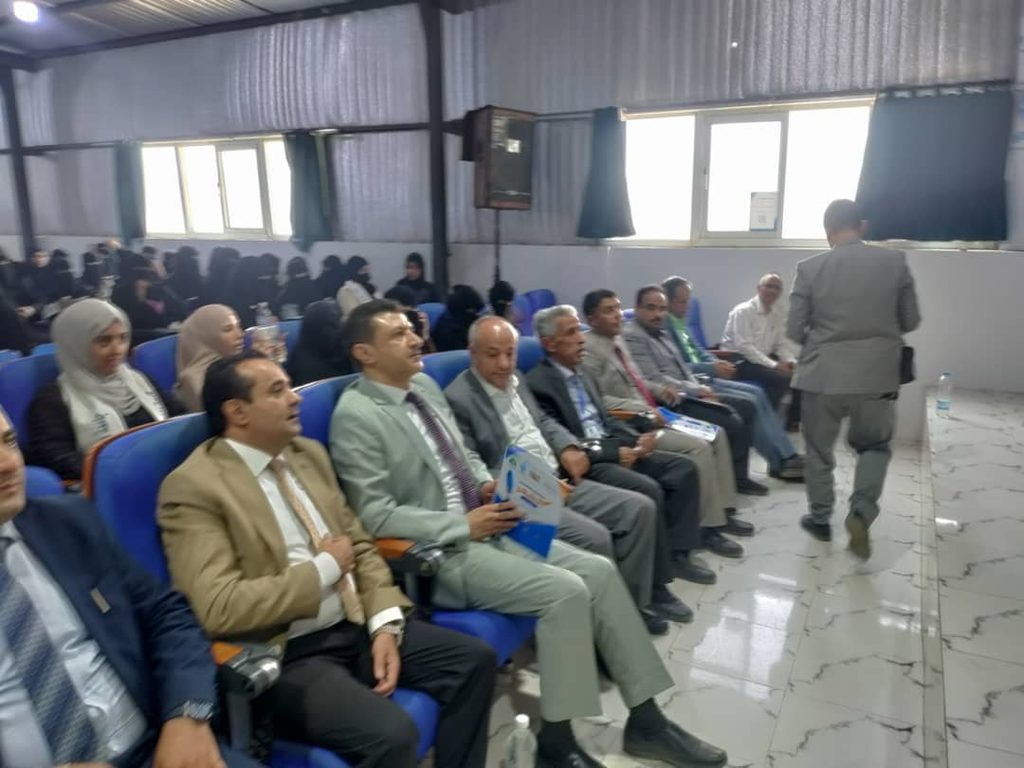Audience seated in a conference hall with rows of blue chairs; men in suits in the foreground and women in hijabs toward the back, as a presenter walks toward the front.