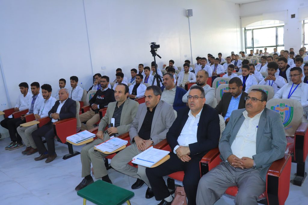 Group of men in a conference hall seated in rows, many wearing white shirts; a camera on a tripod is centered.