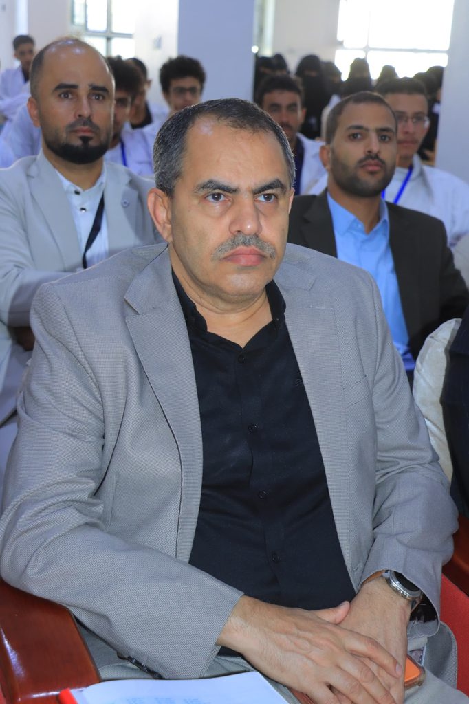 Close-up of a middle-aged man in a gray blazer seated, hands folded, at a conference with other attendees in the background