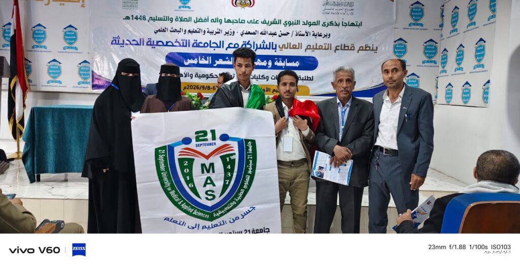Group of people on a stage posing with a banner featuring a shield logo and the text 21 MAS, during an academic event at a university auditorium with a blue-and-white backdrop.