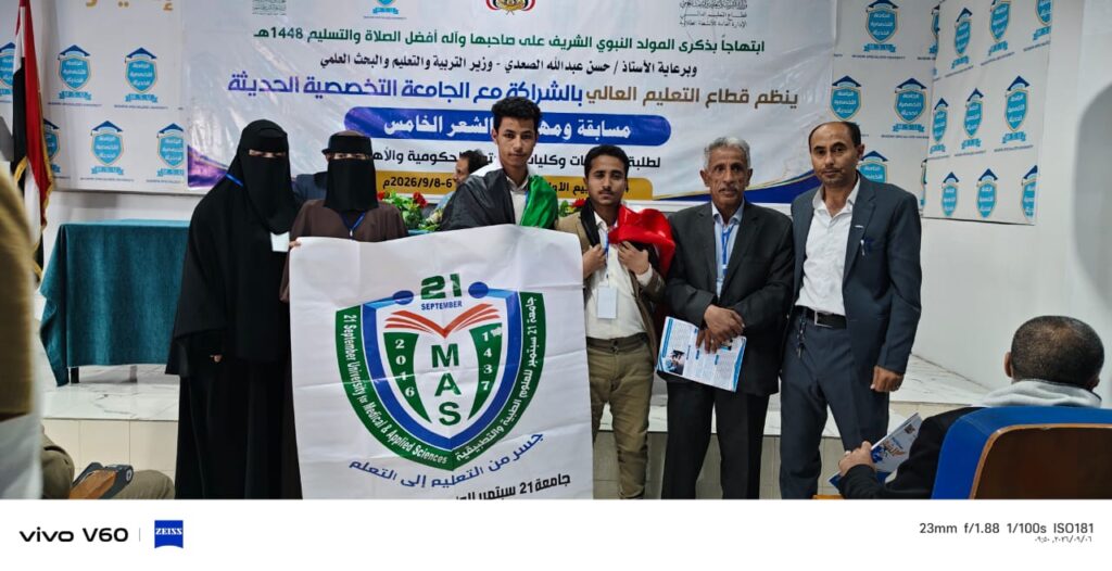 Group of students and officials posing with a MAS banner at a university event or competition room with a large Arabic banner in the background.