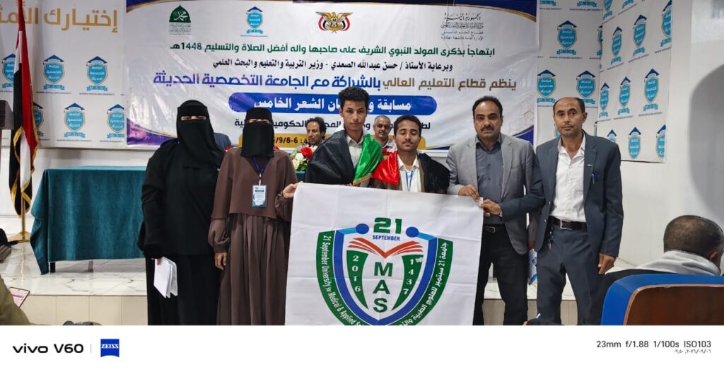 Group of men and women on a stage holding a banner with a green MAS emblem, during an Arabic-language ceremony event.