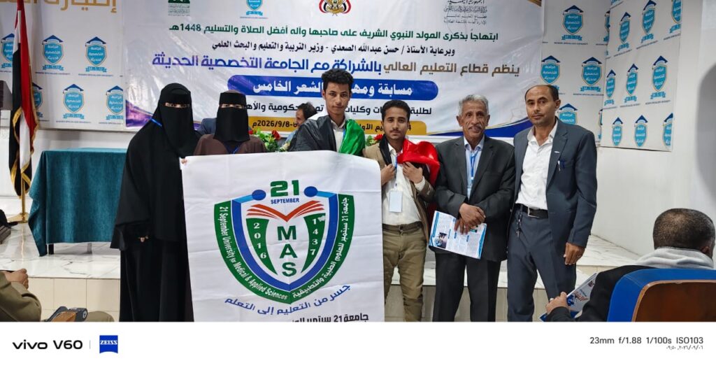 Group of people in a formal event holding a banner with a university emblem and ‘21 MAS’, blue and green shield, in a decorated hall with a large backdrop banner in Arabic.