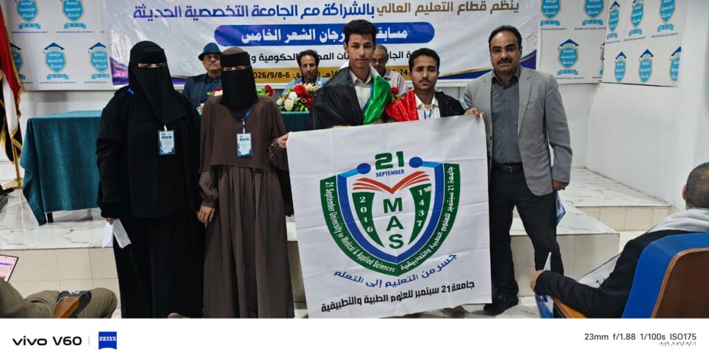 Group of students and officials on a stage holding a large MAS emblem banner at a university event.