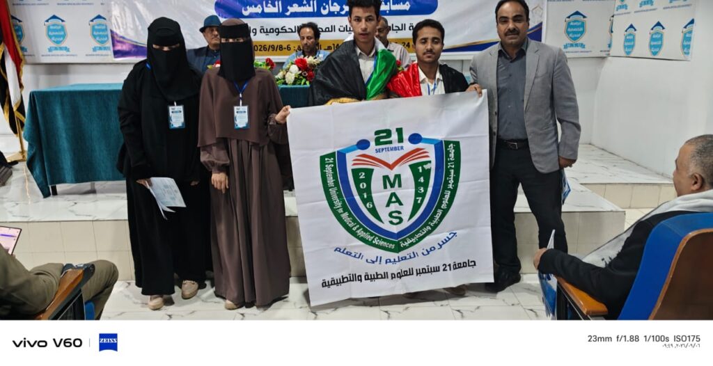 Group of people at a formal event posing with a banner featuring a MAS logo; two women in black niqab stand on the left and men and students stand center-right, banners visible in the background with Arabic text.