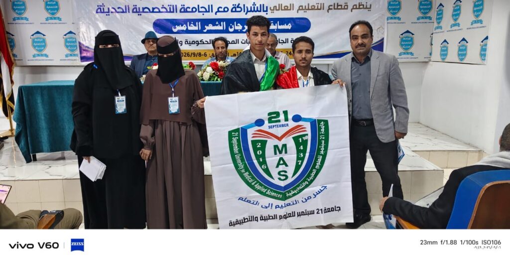 Group of students and a presenter posing indoors, holding a banner with the MAS emblem while a large Arabic banner and logos appear on the wall behind them