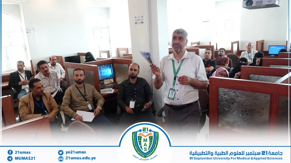 Instructor with a green lanyard presenting to a seated group in a computer lab classroom; attendees listen and take notes.