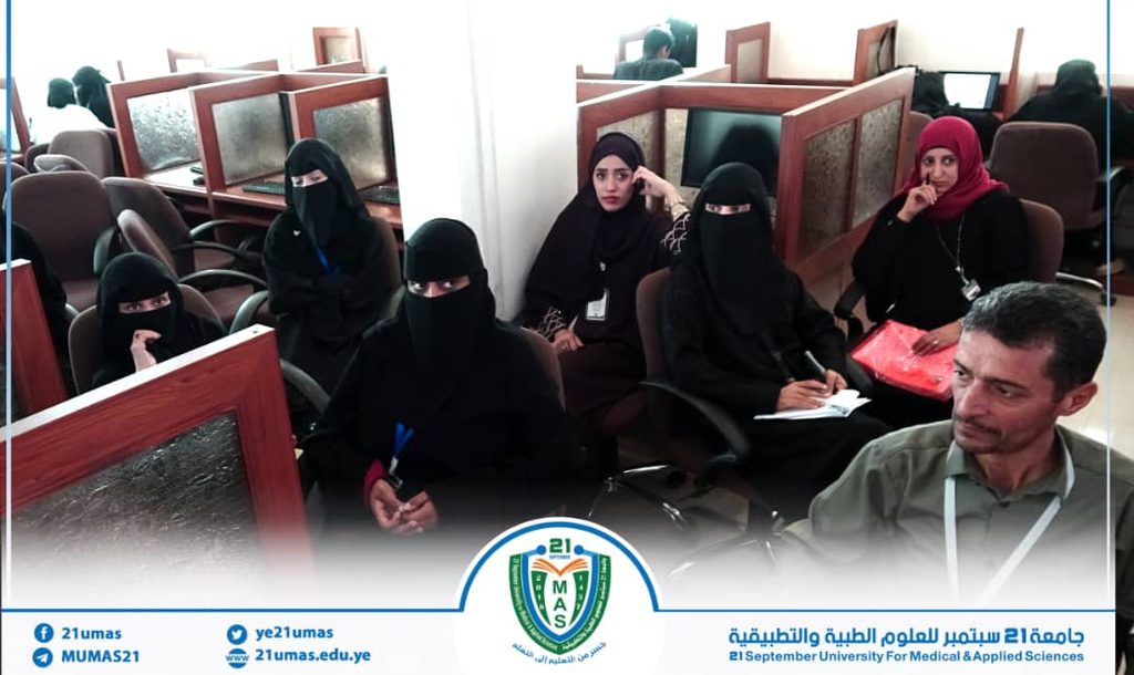 Women wearing hijabs seated in a computer lab with cubicles; a man sits at the right side; university banner at the bottom.