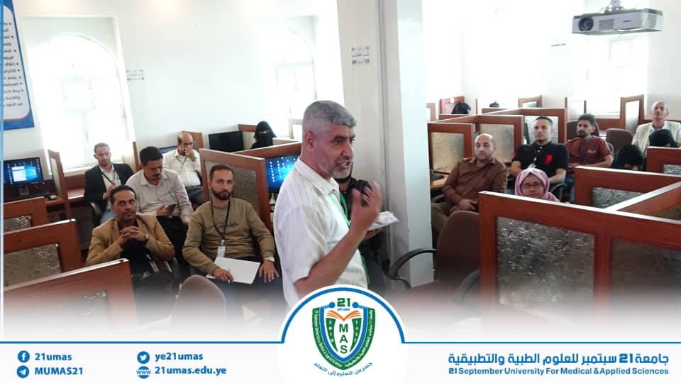 Male instructor speaks to a classroom of adult students seated in individual desks in a seminar room at a university campus.