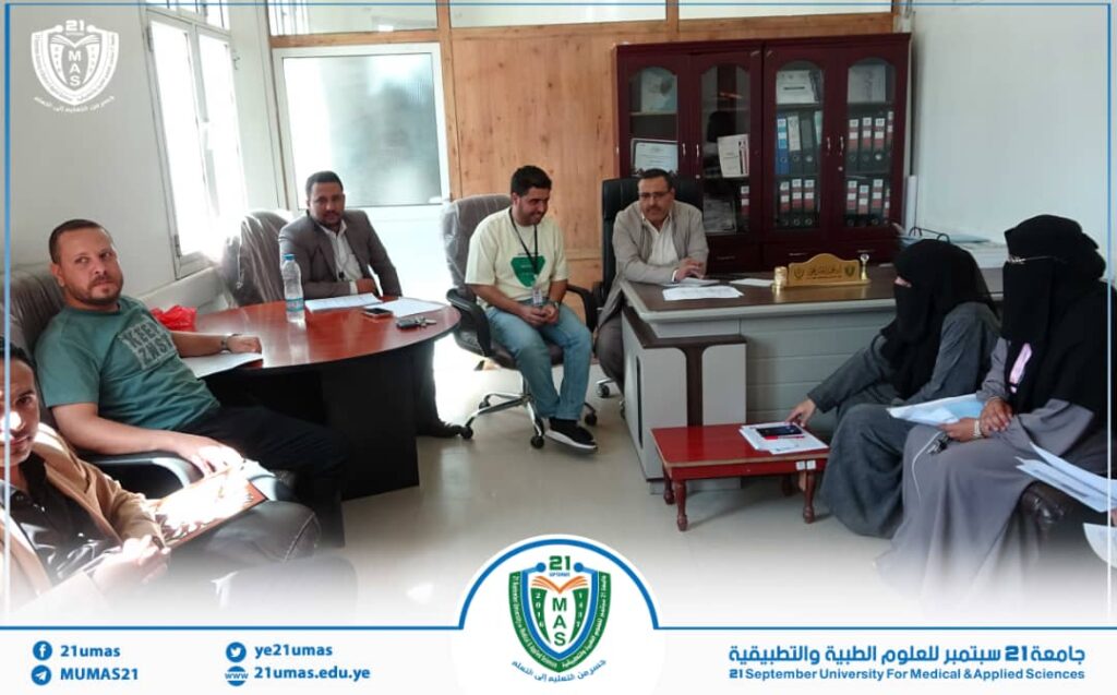 Group of people in a meeting room around a desk at a university office, with a wooden cabinet and logo in corner.