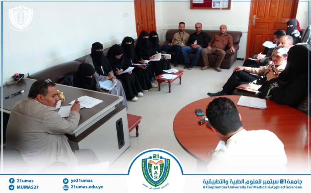 Group of people in a formal university meeting: women in black abayas and men seated around sofas and desks, taking notes with a university logo visible at the bottom.]