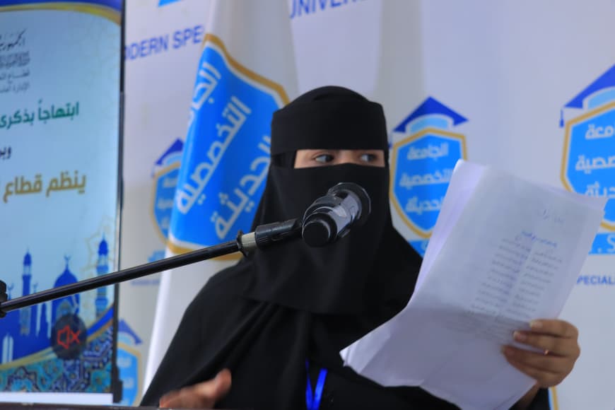 IMG-20260907-WA0035 - جامعة 21 سبتمبر A woman wearing a black niqab speaks into a microphone while holding papers at a podium, with event banners in the background.