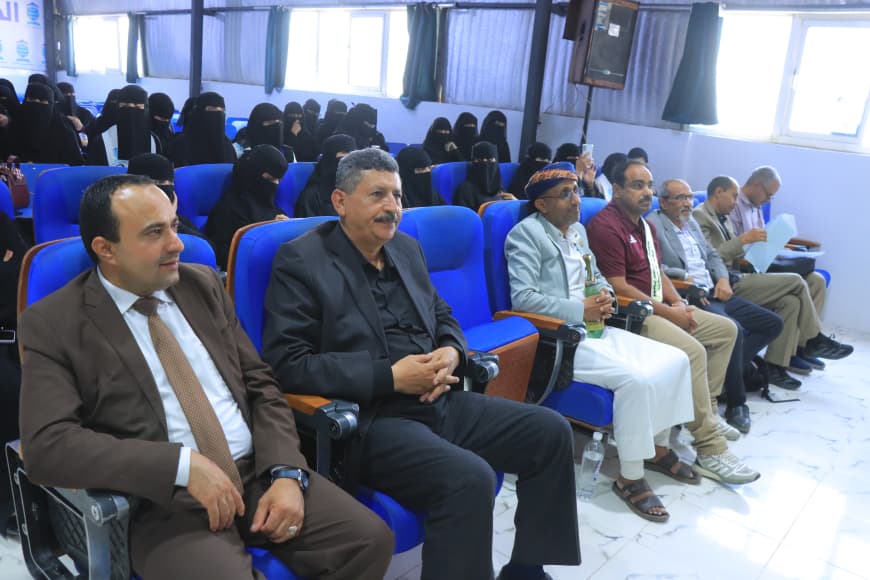 IMG-20260907-WA0036 - جامعة 21 سبتمبر A row of men seated in blue auditorium chairs at a formal event, some wearing suits and traditional clothing. Behind them, women in black niqab sit in the audience.