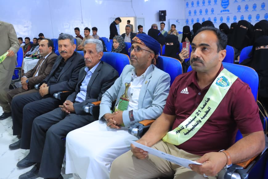 IMG-20260907-WA0037 - جامعة 21 سبتمبر Front row of men in formal attire at an indoor event; man on the far right wears a maroon shirt and light sash, holding papers.
