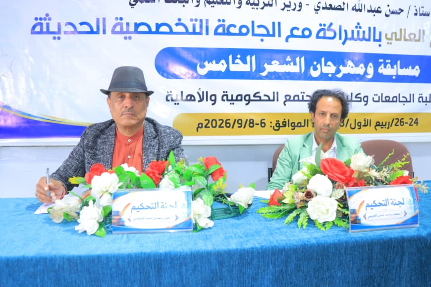 IMG-20260907-WA0038 - جامعة 21 سبتمبر Two men sit at a blue table with flower arrangements at an Arabic event; a large banner behind them advertises a poetry contest, with one man wearing a hat and orange shirt and the other in a green jacket.